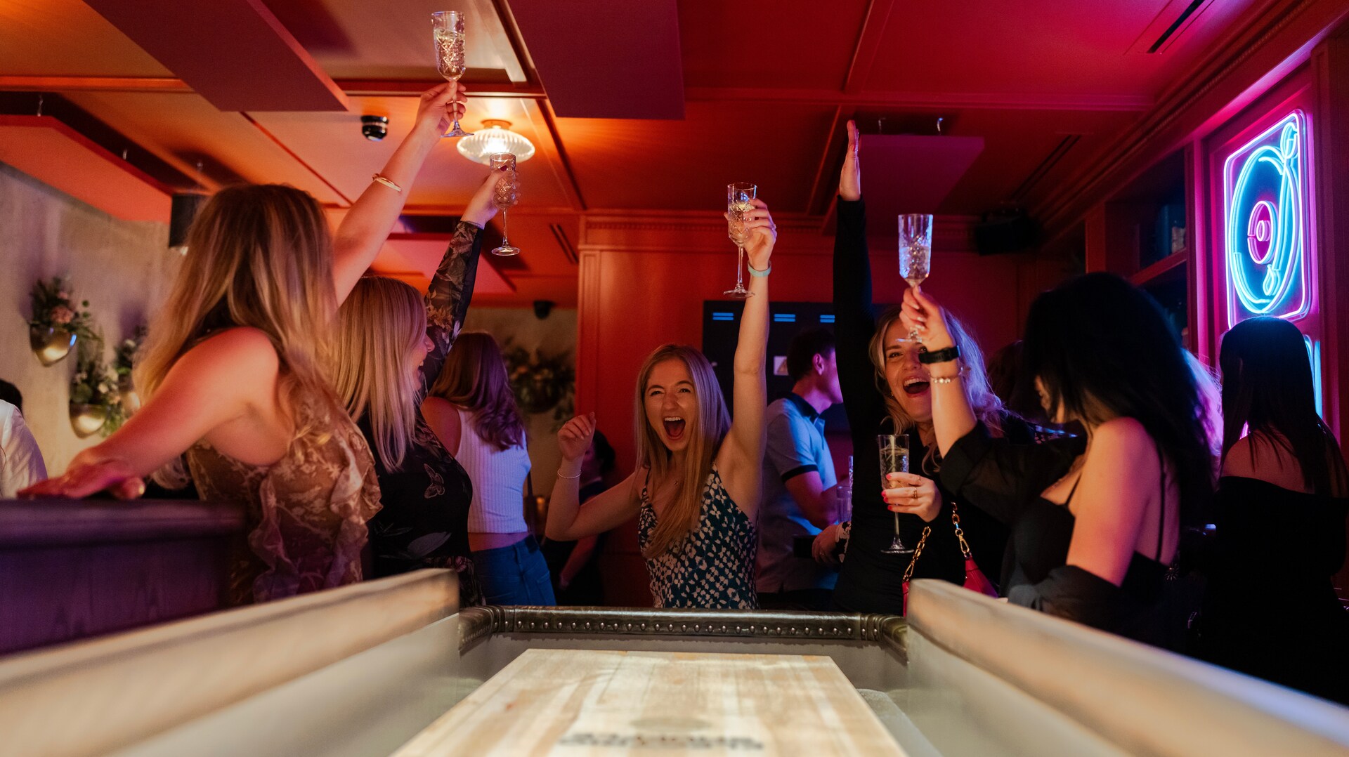 Women celebrating with drinks at a shuffleboard table.